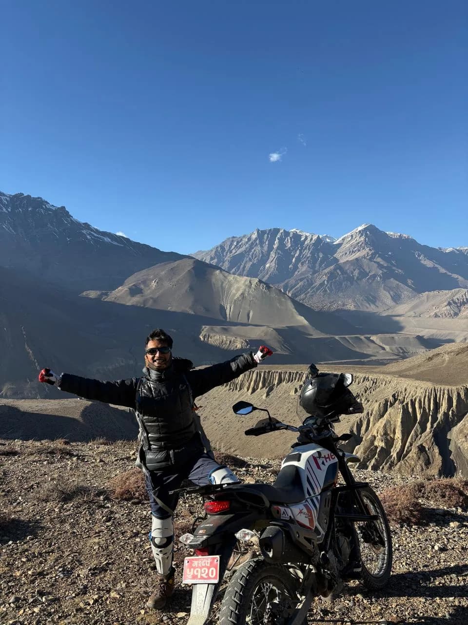 Rohit standing beside his trail bike above the eroded canyon walls of the Kali Gandaki in Mustang, snow peaks on the skyline