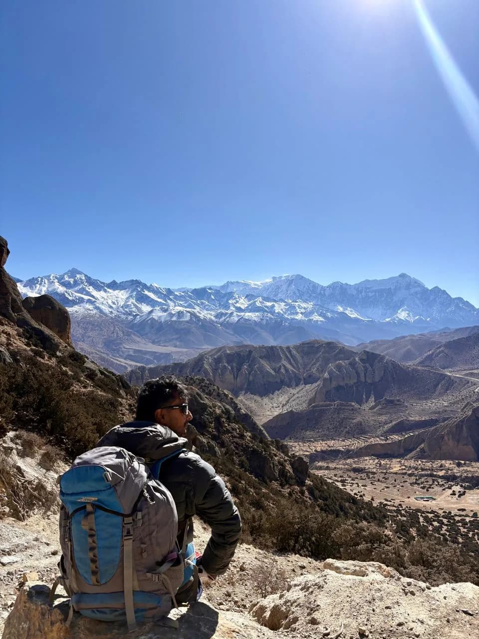 Rohit sitting with his pack at a Mustang viewpoint, looking across the dry valley to a long snow range